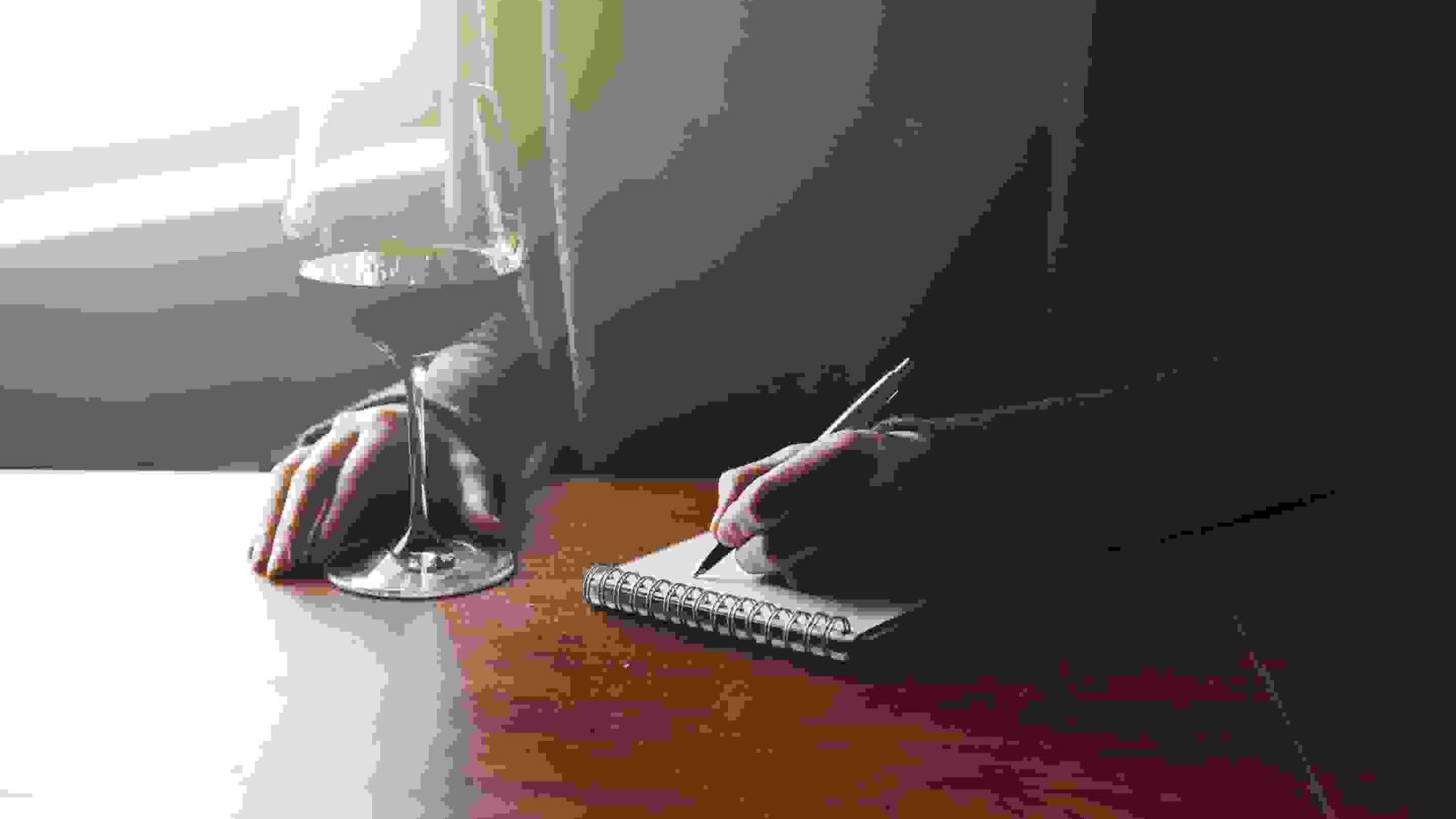 female hands with pen and glass of wine writing on notebook